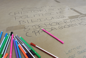 Das Bild zeigt einen Ausschnitt aus einem Tisch auf den Packpapier geklebt wurde. Es ist sind ein Schriftzug und viele bunte Stifte zu sehen und
