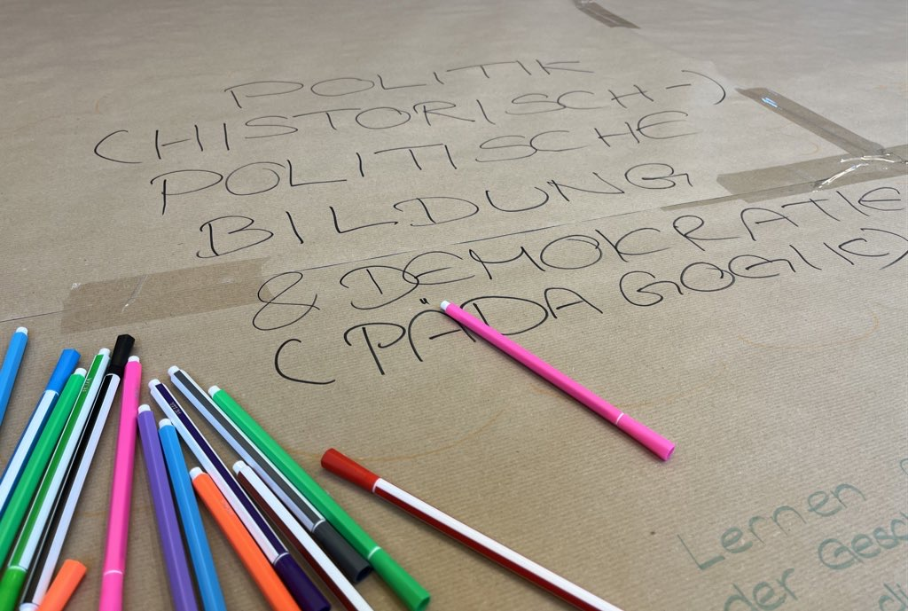 Das Bild zeigt einen Ausschnitt aus einem Tisch auf den Packpapier geklebt wurde. Es ist sind ein Schriftzug und viele bunte Stifte zu sehen und